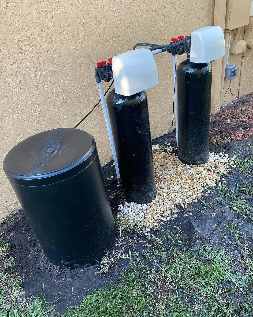 Water softener system with two resin tanks and a brine tank installed on gravel beside a home